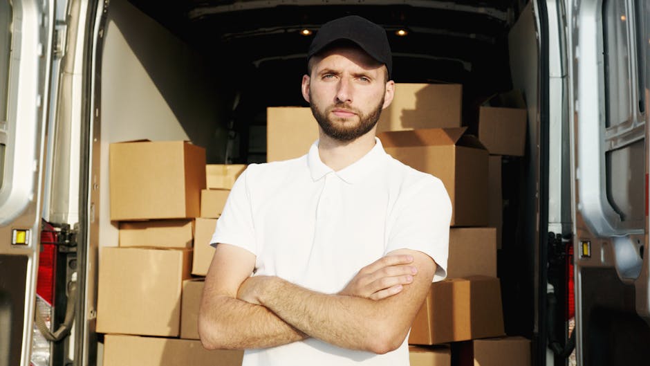 A young man with a serious expression, wearing a white polo shirt and a black cap, stands inside an open moving van filled with numerous stacked brown cardboard boxes of various sizes, some wrapped in