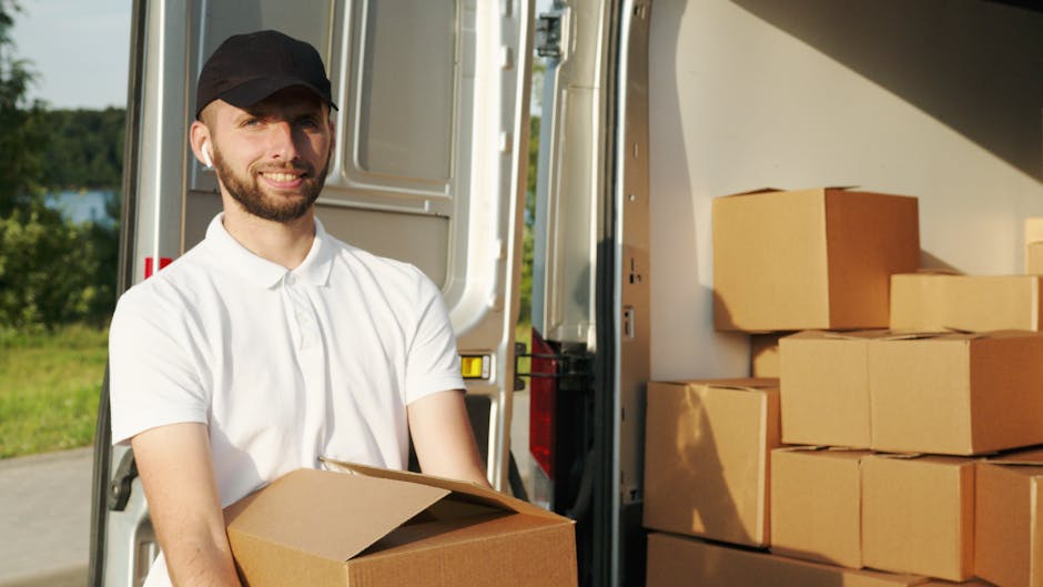 A young man with a serious expression, wearing a white polo shirt and a black cap, stands inside an open moving van filled with numerous stacked brown cardboard boxes of various sizes, some wrapped in