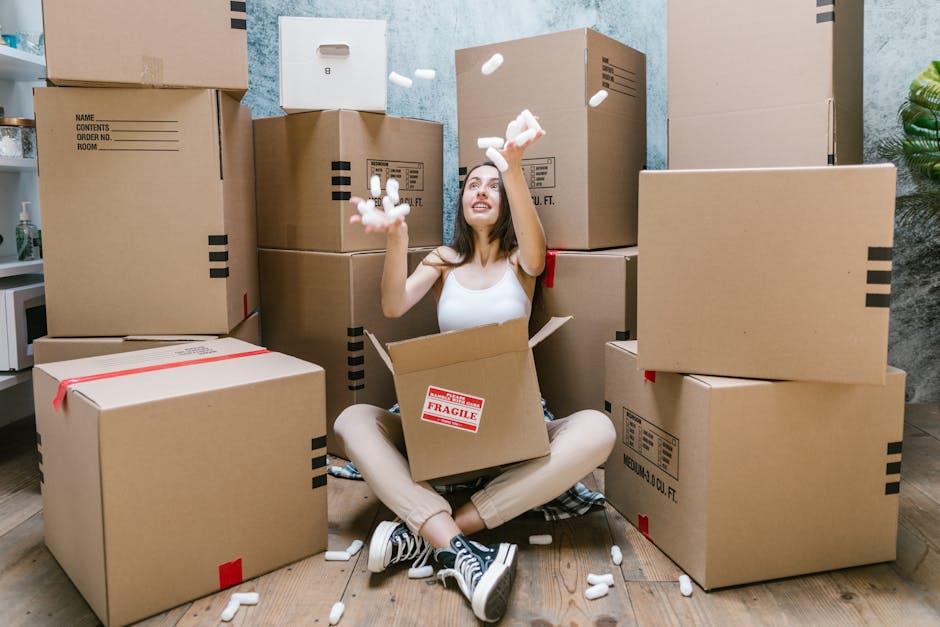 A person wearing a grey t-shirt is carrying a large, plain brown cardboard box at a house interior. The box has flaps open at the top and is being held with one hand supporting the bottom. Behind, par