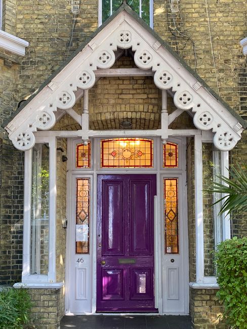 The image displays the entrance of a house with a prominent Victorian-style porch featuring decorative white timber detailing and a gabled roof. The front door is painted a vivid purple, contrasting w